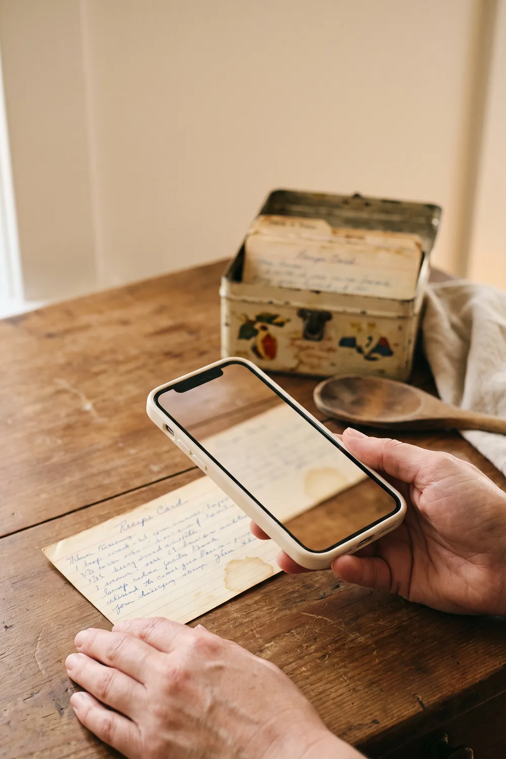Hands holding a phone above a stained handwritten recipe card on a wooden table, with a tin box of more recipe cards and a wooden spoon behind it